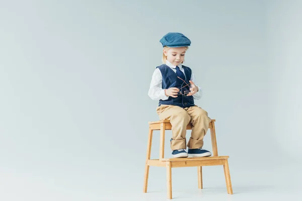 Adorable enfant assis sur des escaliers en bois et tenant des lunettes sur fond gris — Photo de stock