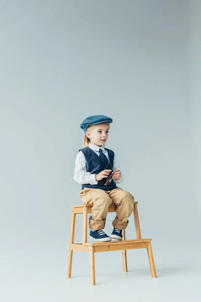 Adorable kid sitting on wooden stairs, holding glasses and looking away on grey background — Stock Photo