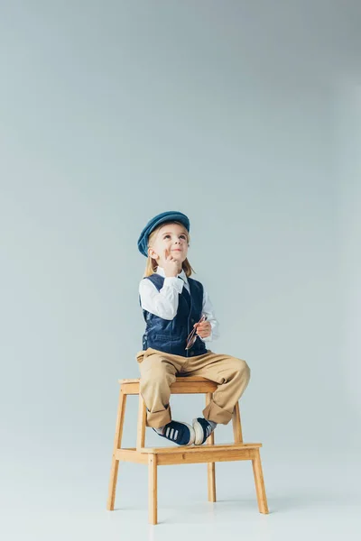Garoto pensativo sentado em escadas de madeira e segurando óculos no fundo cinza — Fotografia de Stock