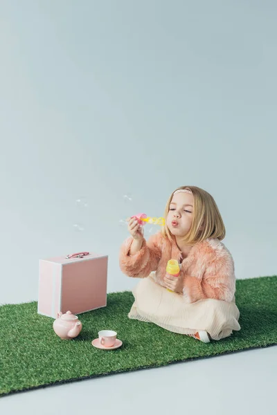 Mignon enfant avec les jambes croisées assis sur tapis d'herbe et soufflant bulles de savon isolé sur gris — Photo de stock