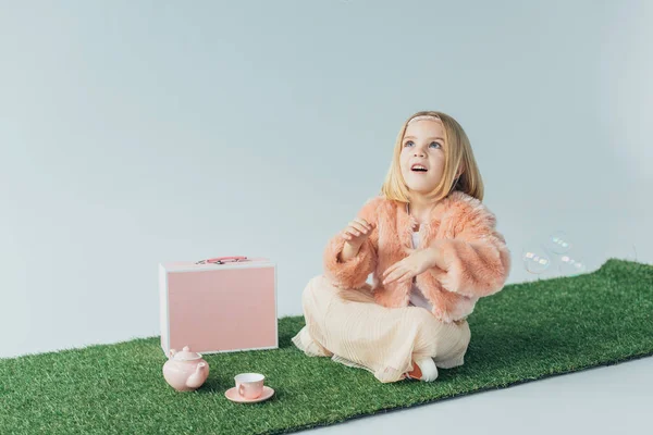 Mignon enfant avec les jambes croisées assis sur tapis en herbe et en regardant des bulles de savon isolé sur gris — Photo de stock