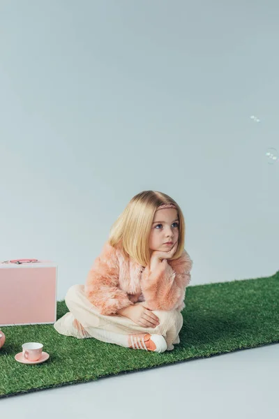 Niño reflexivo con las piernas cruzadas sentado en la alfombra de hierba y mirando hacia otro lado aislado en gris - foto de stock