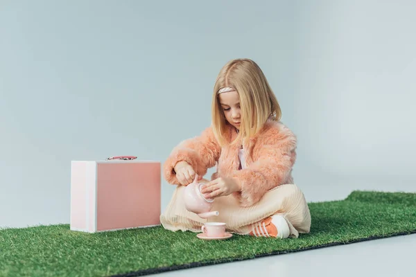 Niño sentado con las piernas cruzadas jugando con platos de juguete rosa aislado en gris - foto de stock