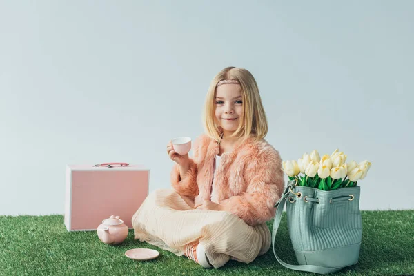 Lächelndes Kind mit überkreuzten Beinen, das mit Spielzeuggeschirr spielt und isoliert auf grau in die Kamera schaut — Stockfoto