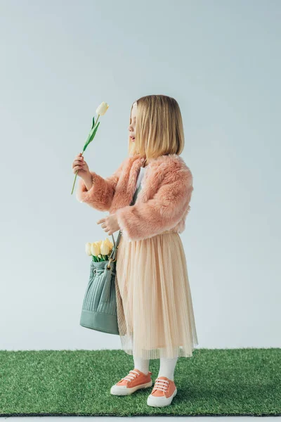 Niño sorprendido y lindo con bolsa mirando tulipán blanco aislado en gris - foto de stock