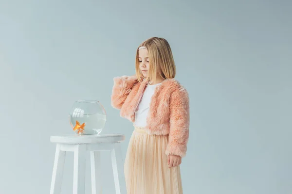 Cute kid in faux fur coat and skirt looking at fishbowl on stool isolated on grey — Stock Photo