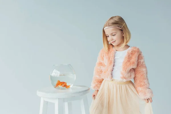 Adorable kid in faux fur coat and skirt looking at fishbowl on stool isolated on grey — Stock Photo
