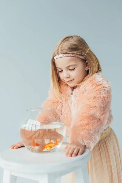 Criança adorável em casaco de pele falso e saia olhando para aquário em banquinho isolado em cinza — Fotografia de Stock