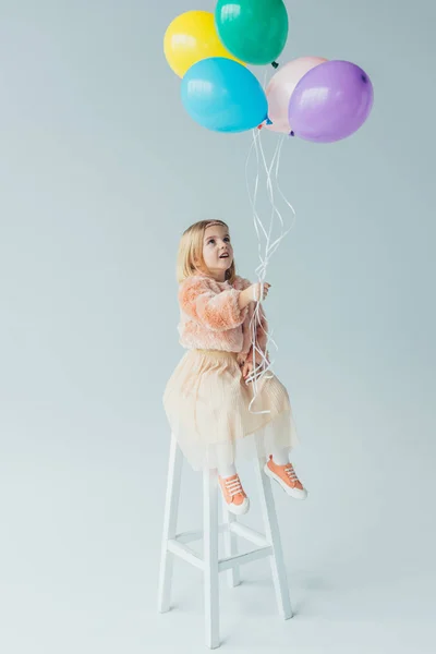 Niedliches Kind in Kunstpelzmantel und Rock sitzt auf Hochstuhl und hält Luftballons — Stockfoto