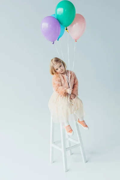 Cute kid in faux fur coat and skirt sitting on highchair, looking at camera and holding balloons — Stock Photo