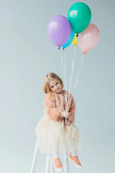 Очаровательный ребенок в искусственной шубе и юбке сидит на стульчике, смотрит в камеру и держит воздушные шары, изолированные на сером — стоковое фото