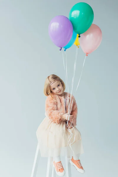 Adorable niño en abrigo de piel sintética y falda sentado en la trona, mirando a la cámara y sosteniendo globos aislados en gris — Stock Photo