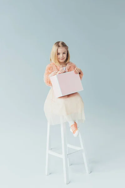 Ребенок в искусственном шубе и юбке сидит на стульчике и держит розовый чехол на сером фоне — стоковое фото