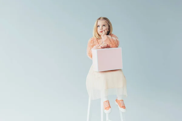 Enfant réfléchi en fausse fourrure manteau et jupe assis sur chaise haute et tenant le cas rose isolé sur gris — Photo de stock