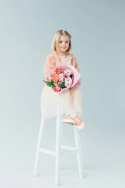 Kid in faux fur coat and skirt sitting on highchair and holding bouquet on grey background — Stock Photo
