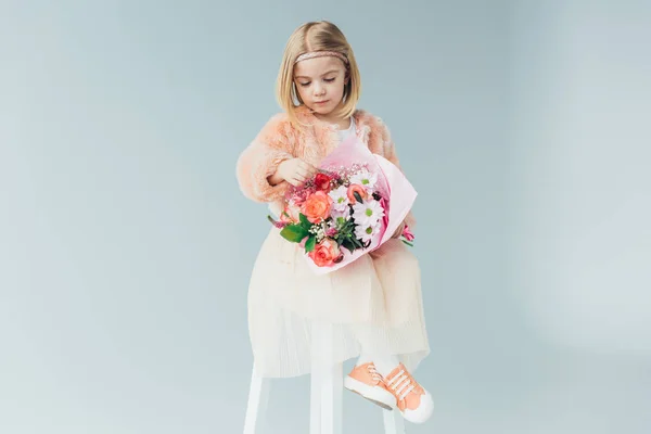 Cute kid in faux fur coat and skirt sitting on highchair and holding bouquet isolated on grey — Stock Photo