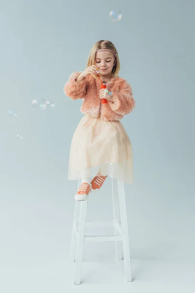 Adorable kid in faux fur coat and skirt sitting on highchair and blowing soap bubbles — Stock Photo