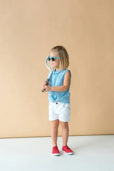 Cute kid in shirt and shorts holding racket on beige background — Stock Photo