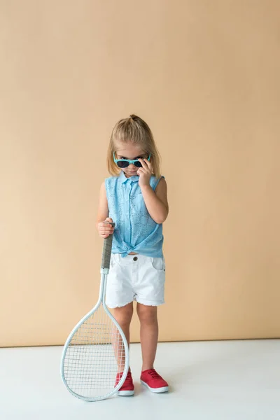 Cute kid in shirt and shorts holding racket and looking at camera on beige background — Stock Photo