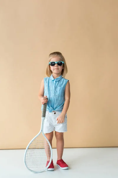 Cute kid in sunglasses, shirt and shorts holding racket and looking at camera — Stock Photo