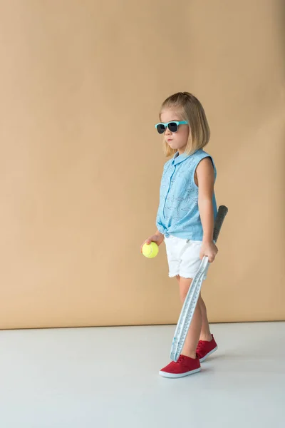 Mignon enfant en lunettes de soleil, chemise et short tenant raquette et ballon sur fond beige — Photo de stock