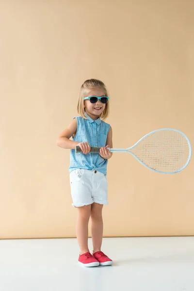 Smiling and cute kid in sunglasses, shirt and shorts holding racket on beige background — Stock Photo