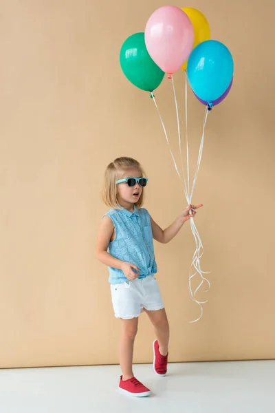 Cute kid in sunglasses, shirt and shorts holding balloons on beige background — Stock Photo