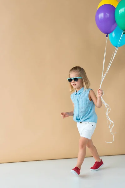 Lindo niño en gafas de sol, camisa y pantalones cortos caminando y sosteniendo globos - foto de stock
