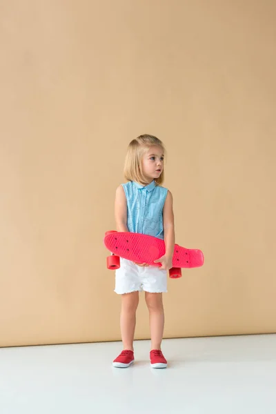 Cute kid in shirt and shorts holding pink penny board on beige background — Stock Photo