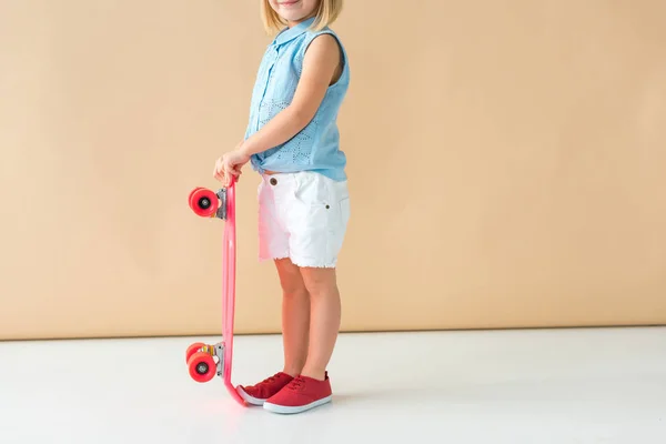 Partial view of kid in shirt and shorts holding pink penny board on beige background — Stock Photo