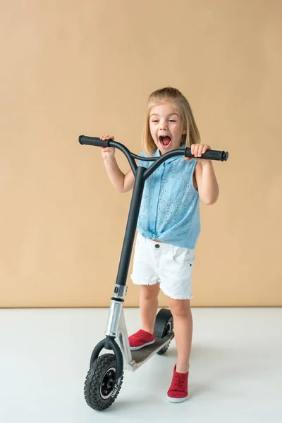 Bambino sorridente in camicia e pantaloncini equitazione scooter su sfondo beige — Foto stock
