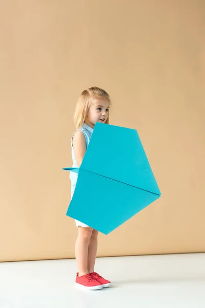 Criança adorável brincando com avião de papel azul no fundo bege — Fotografia de Stock