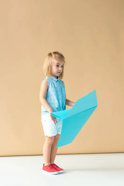 Surprised  kid in shirt and shorts holding blue paper plane on beige background — Stock Photo