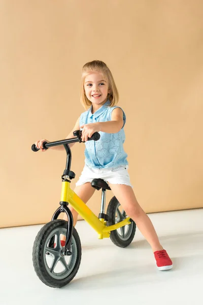 Niño sonriente en camisa y pantalones cortos montar en bicicleta sobre fondo beige - foto de stock