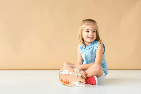 Cute kid sitting on floor with fishbowl and looking away on beige background — Stock Photo