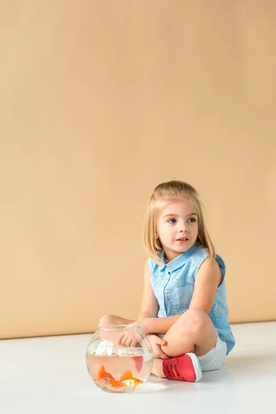 Niño pensativo sentado en el suelo con pecera y mirando hacia otro lado sobre fondo beige - foto de stock