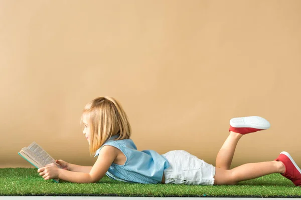 Side view of kid lying on grass rug and reading book on beige background — Stock Photo
