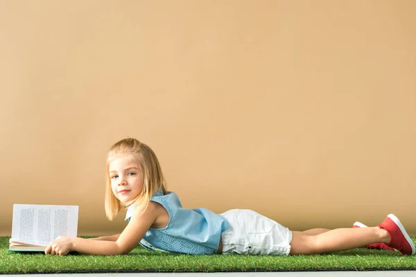 Cute kid lying on grass rug and holding book on beige background — Stock Photo