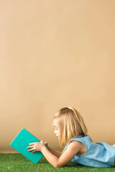 Kid lying on grass rug and reading book on beige background — Stock Photo