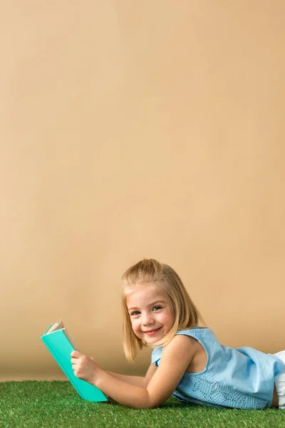Sonriente y lindo niño acostado en la alfombra de hierba y sosteniendo libro sobre fondo beige - foto de stock