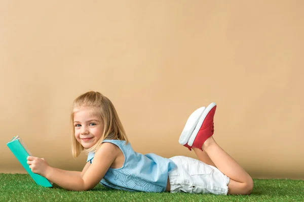 Enfant souriant et mignon couché sur tapis en herbe et tenant livre sur fond beige — Photo de stock