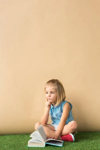 Enfant coûteux assis avec les jambes croisées sur tapis en herbe et tenant livre sur fond beige — Photo de stock