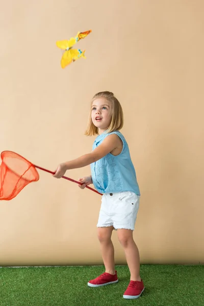 Cute kid catching butterfly with red butterfly net on beige background — Stock Photo