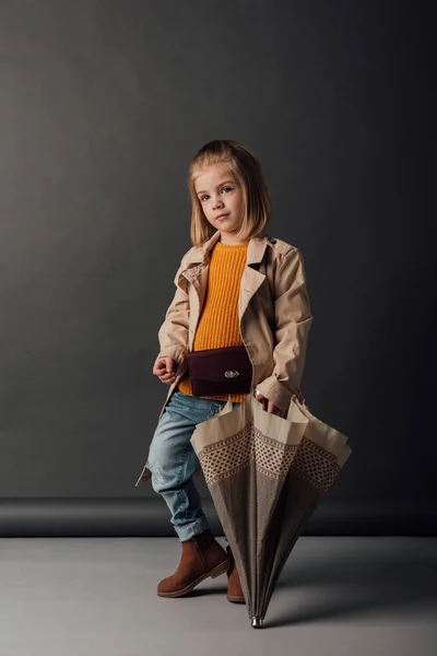 Kid in trench coat and jeans holding umbrella and looking away — Stock Photo