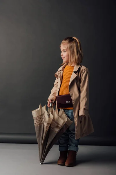 Cute kid in trench coat and jeans holding umbrella and looking away — Stock Photo