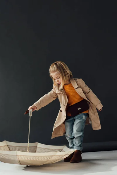 Child in trench coat and jeans playing with umbrella on black background — Stock Photo