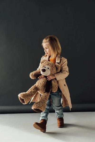 Cute child in trench coat and jeans holding teddy bear on black background — Stock Photo