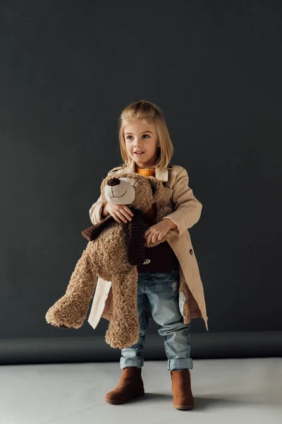 Niño sonriente y lindo en gabardina y jeans sosteniendo oso de peluche sobre fondo negro - foto de stock