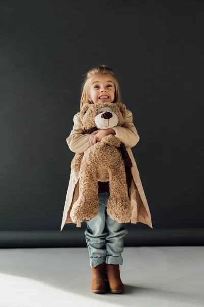 Criança feliz e bonito em casaco de trincheira e jeans segurando ursinho de pelúcia no fundo preto — Fotografia de Stock