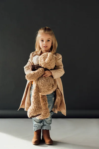 Überrascht und niedlich Kind in Trenchcoat und Jeans hält Teddybär auf schwarzem Hintergrund — Stockfoto